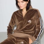 Una mujer con el pelo liso y oscuro está sentada en una silla y lleva puesto un chándal de terciopelo Ralph Velour Pants Brown (copia), con rayas blancas y negras en las mangas y las piernas y un logotipo blanco en el pecho y los pantalones.